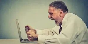 A frustrated man sitting at a desk, clenching fists near a laptop, expressing anger and struggle while working alone.