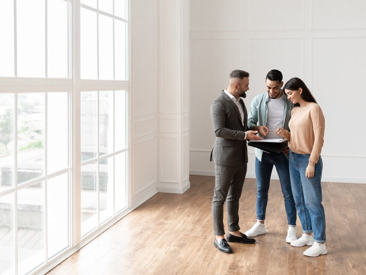 Three people standing in an empty, well-lit room reviewing documents together, representing clients receiving information in a property interior.
