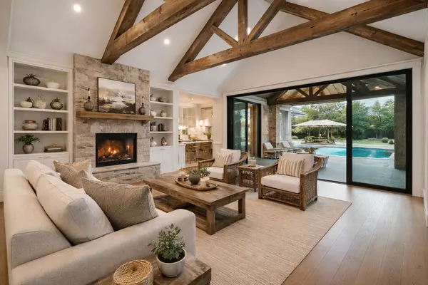 Bright, open-concept living room in a modern farmhouse home featuring vaulted ceilings with exposed wood beams, a stone fireplace, neutral furnishings, and large sliding glass doors opening to a landscaped backyard and pool.
