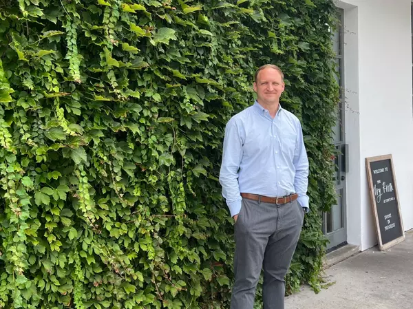Professional real estate advisor standing outdoors beside a lush green living wall, wearing a light blue dress shirt and gray slacks, in a clean, modern commercial setting with natural light.