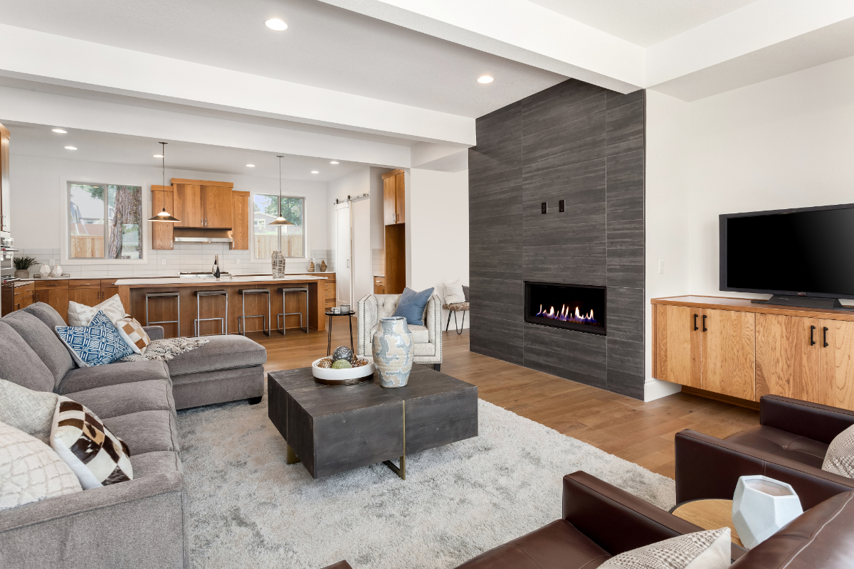 Open-concept living room with kitchen in background, fireplace feature wall to the right, light wood cabinets and grey sofa, wide shot showing seating area and dining island.