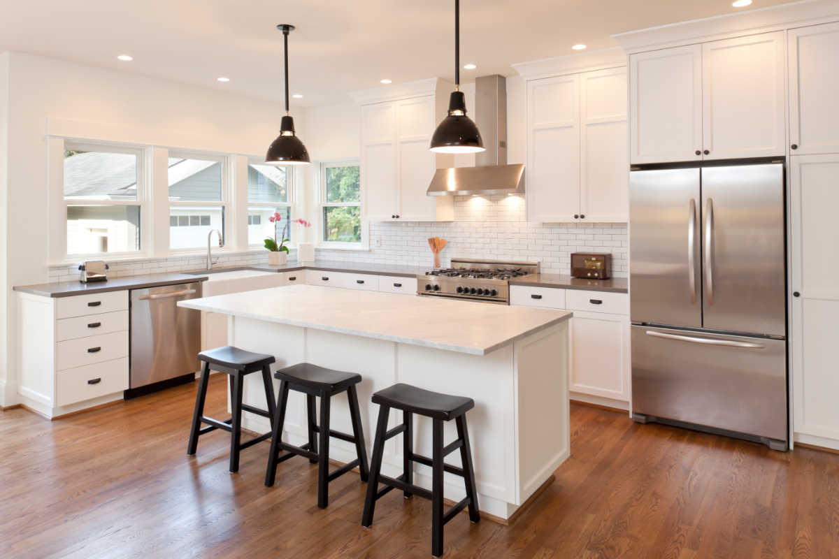 Bright white kitchen with central island and pendant lighting, stools at counter and stainless steel appliances visible.