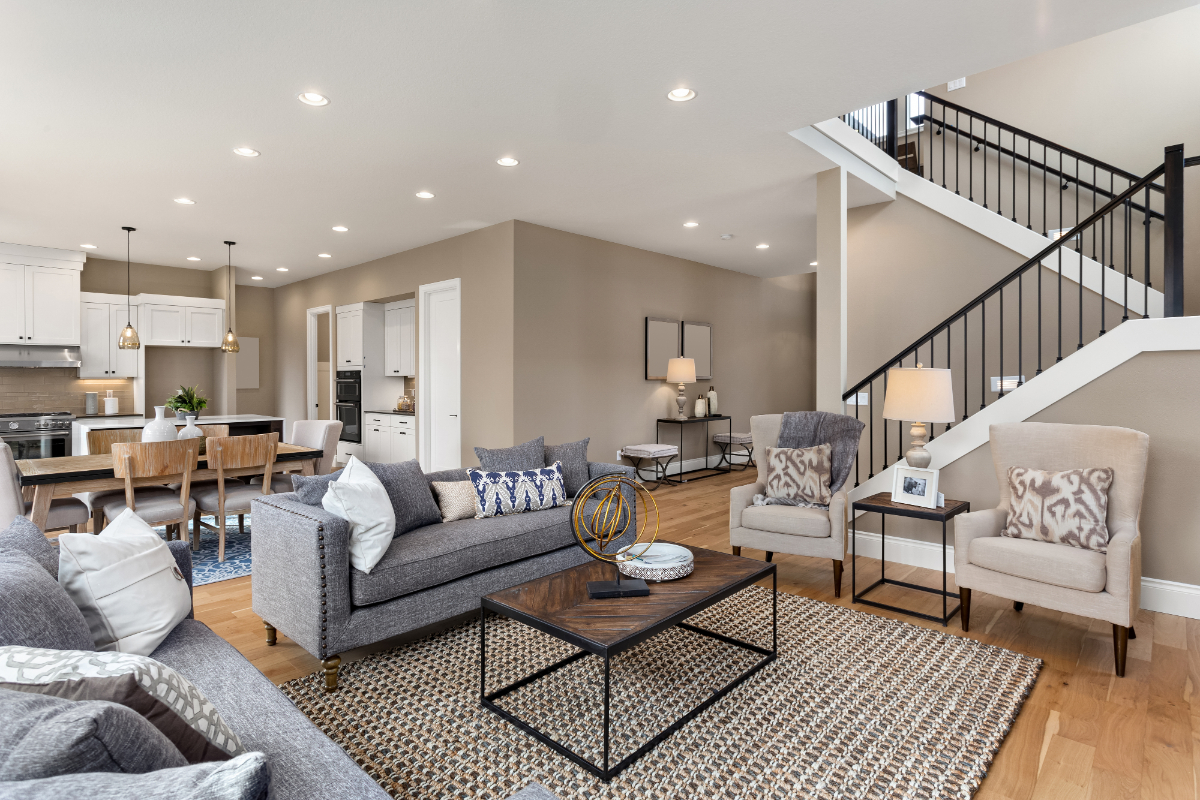 Spacious modern family room and kitchen area, open floor plan with neutral furnishings, staircase visible on right, contemporary lighting and decor.
