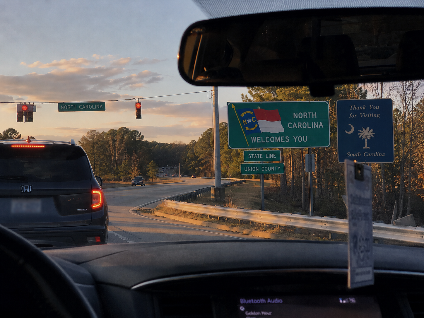 View from inside a car stopped at a traffic light near the North Carolina–South Carolina state line during golden hour. Through the windshield, a large "North Carolina Welcomes You" sign stands beside the road, with a smaller "Union County" sign below it and a "Thank You for Visiting South Carolina" sign nearby. Warm late afternoon sunlight and tree-lined roads create the feeling of frequently crossing between the two states.