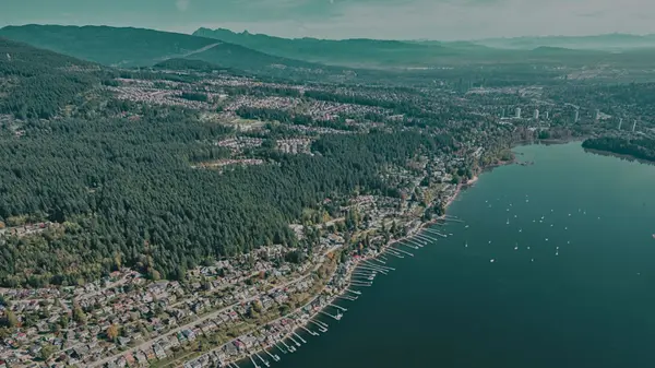 Aerial view of Port Moody’s waterfront, with shoreline homes, docks and boats along Burrard Inlet beneath forested mountains.