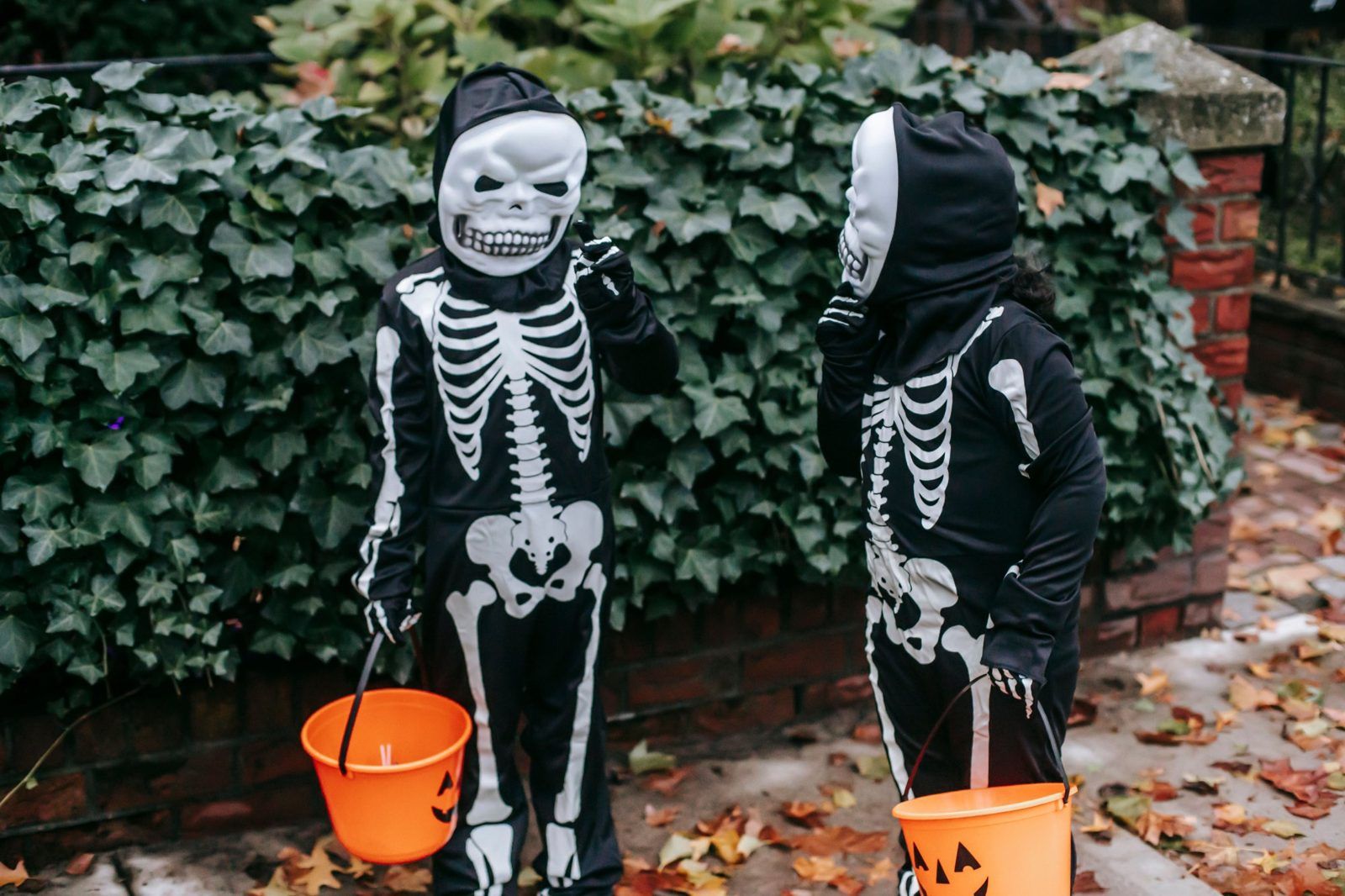 Two kids in skeleton costumes holding jack-o'-lantern candy buckets while trick-or-treating