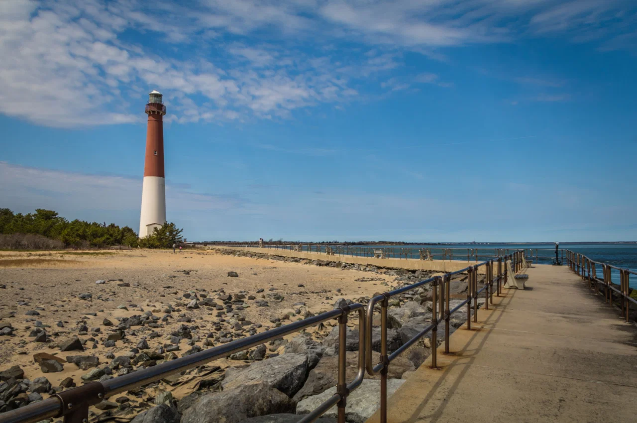 A long concrete pier leading to a tall lighthouse on a rocky shoreline under a blue sky with scattered clouds.