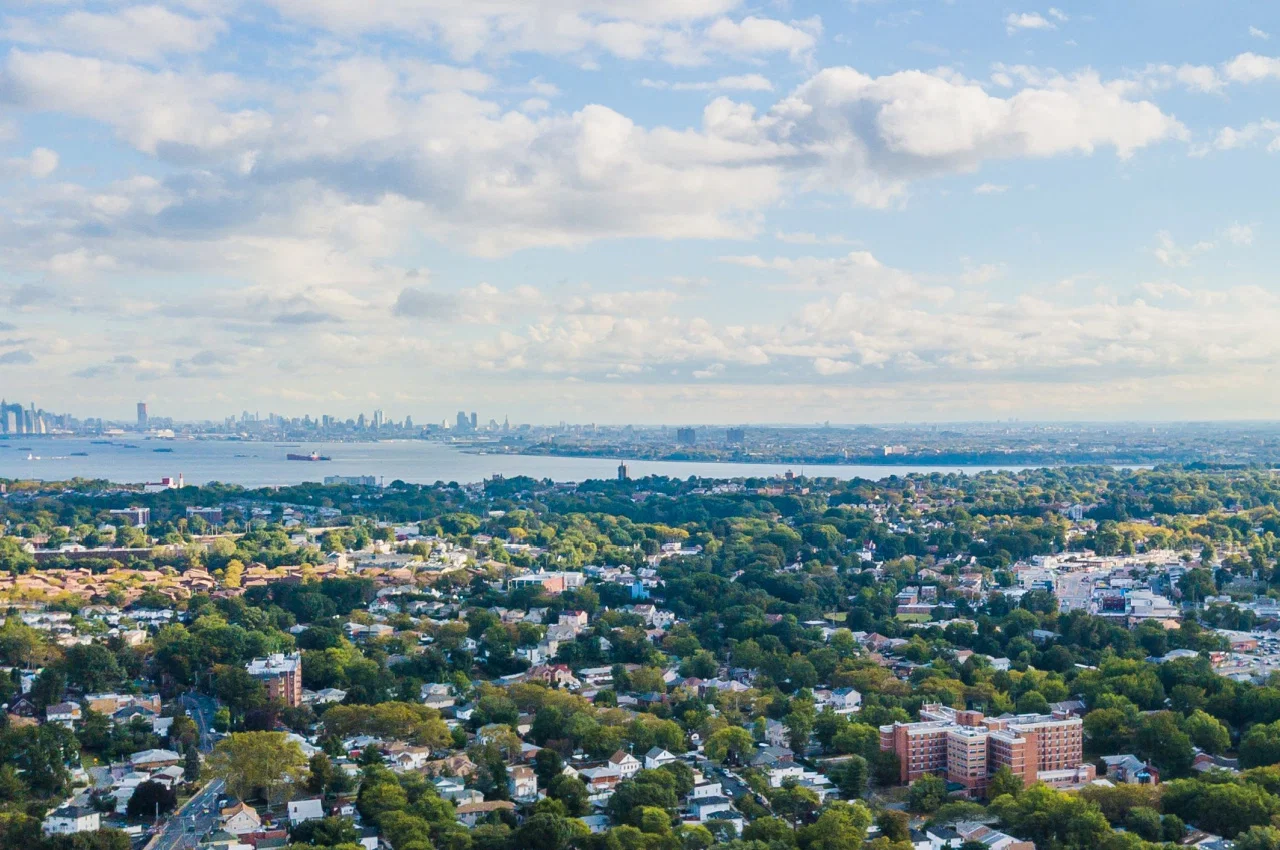 Aerial view of Staten Island showcasing dense residential neighborhoods, trees and distant skyline under a partly cloudy sky.