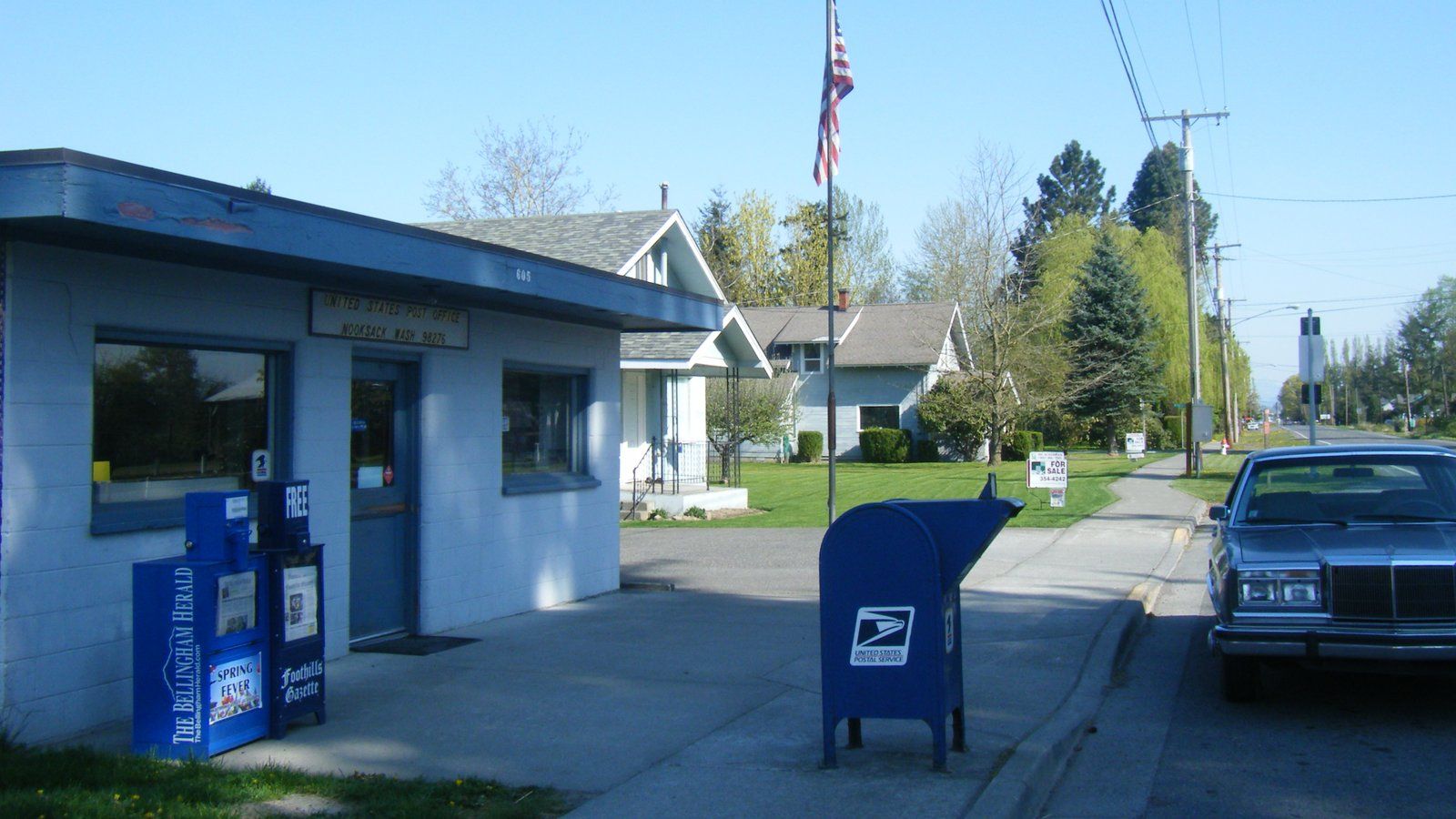 The small blue Nooksack post office, with a flag and a mailbox out front, on a quiet street of older homes in Nooksack.
