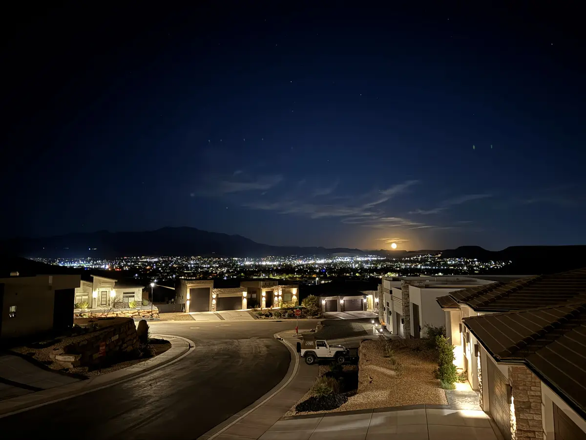 High-angle night view of a quiet St. George neighborhood under a full moon, with glowing streetlights and a sprawling valley of city lights nestled against dark mountain peaks.