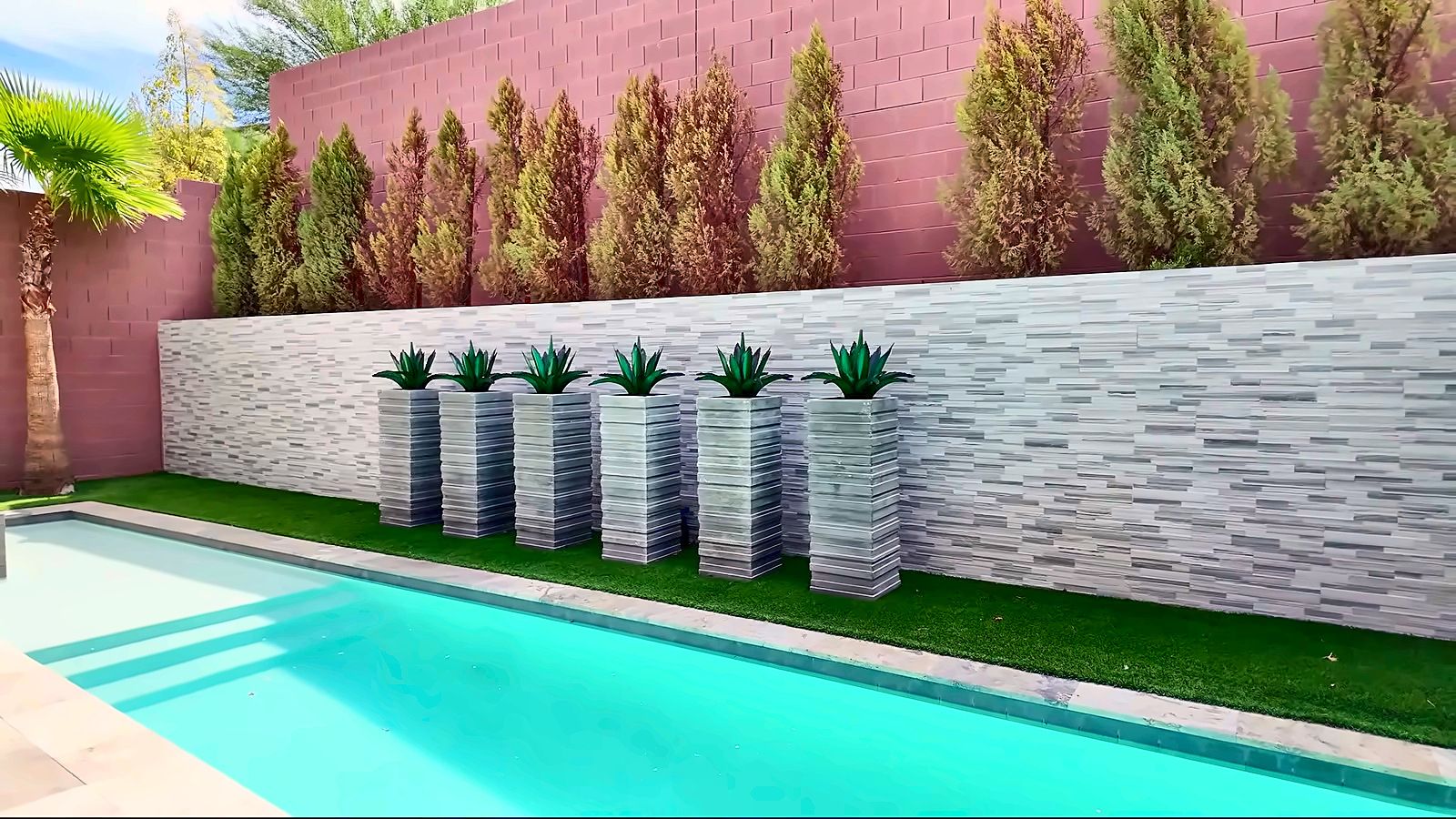 Rectangular backyard pool with clear blue water, a stone-clad wall with planters and a strip of green lawn
