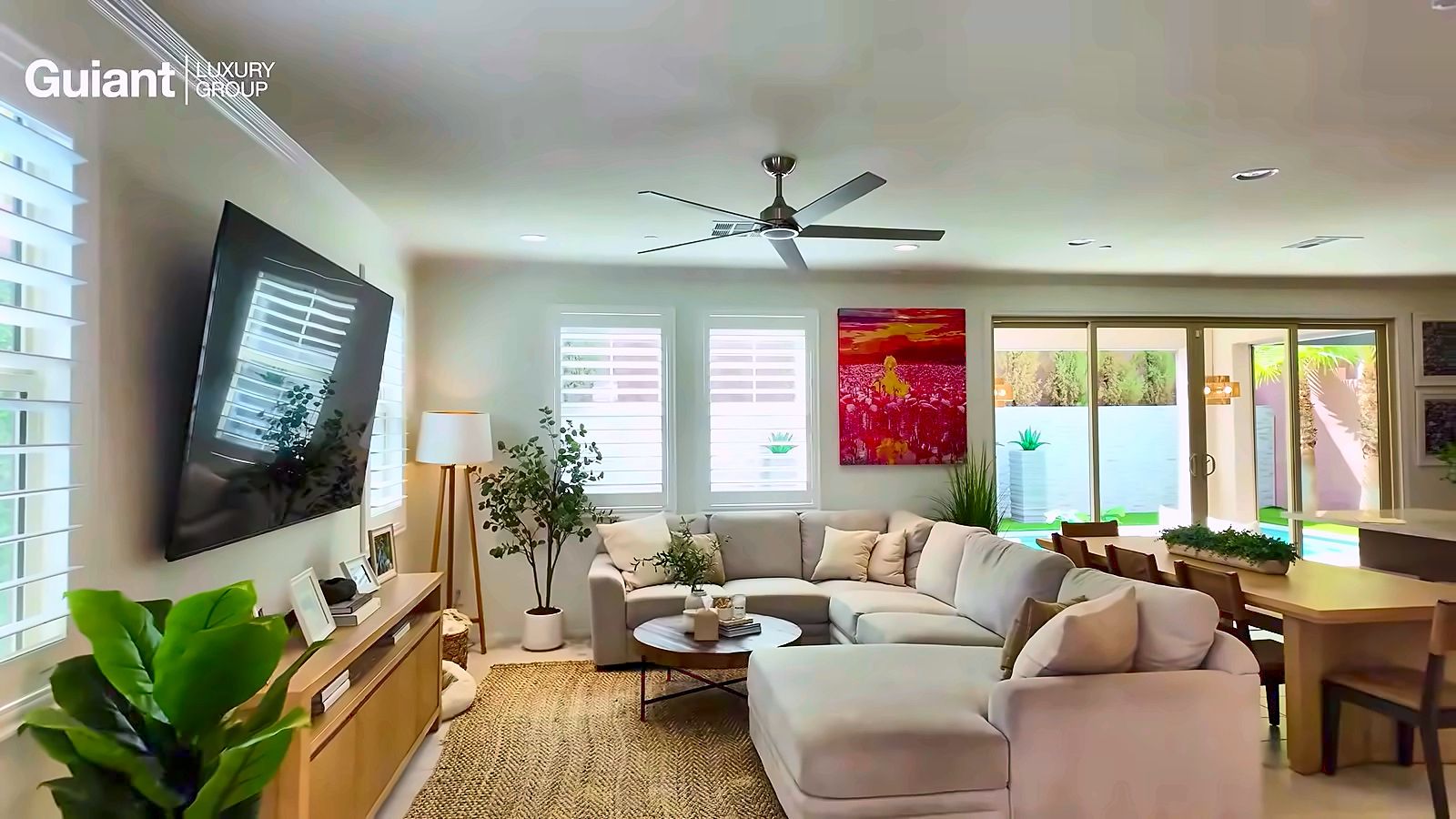 Bright living room with a light sectional, plants, floor lamp and shutters open, flowing into a dining area and backyard slider