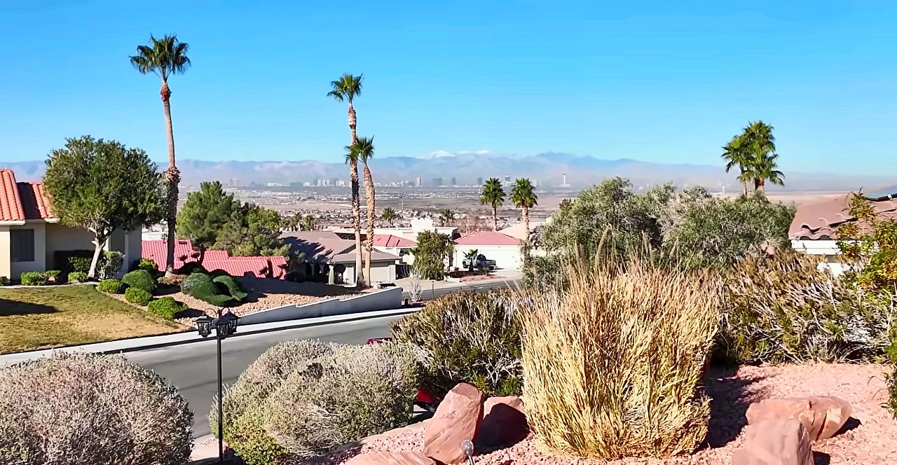 Las Vegas Strip skyline and snow-capped Spring Mountains seen from a Henderson hillside with desert landscaping and palm trees