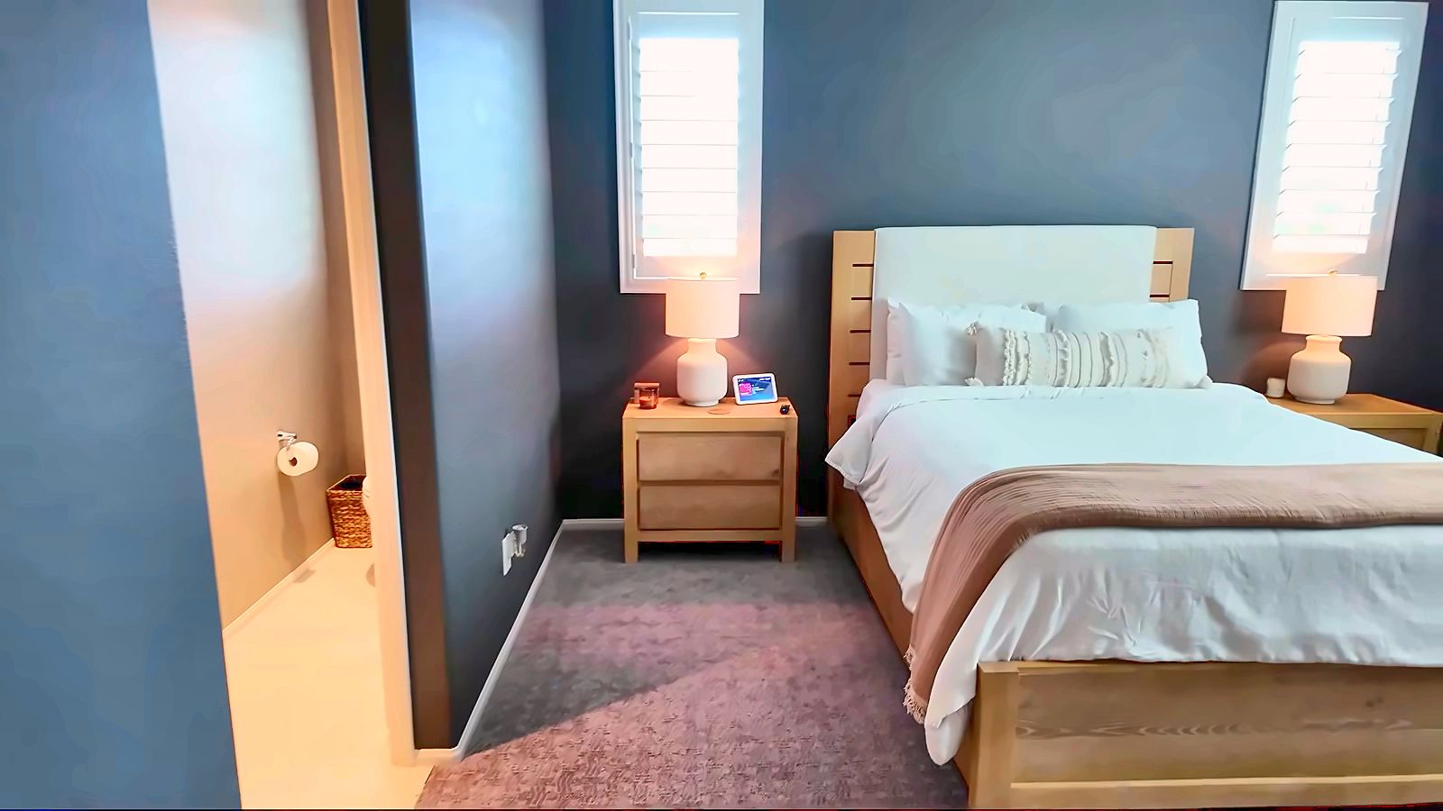 Bedroom with a made bed, light bedding, matching nightstands with both lamps on and white shutters