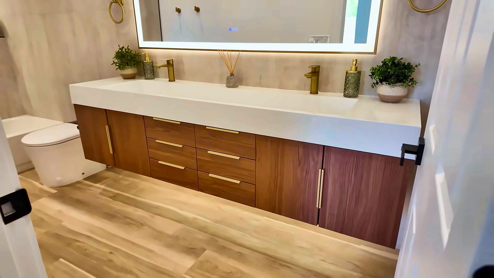 Bathroom with a long white floating vanity, wood cabinets, gold faucets, a lit mirror and only a plant and soap on the counter