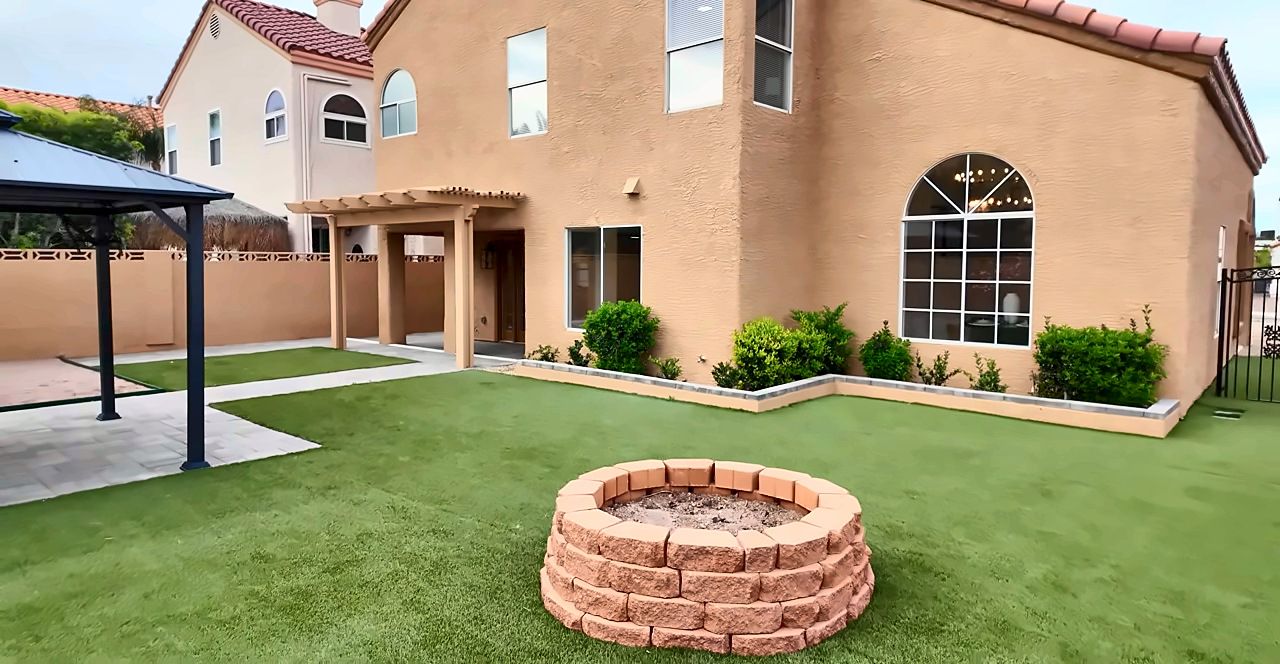 Backyard with artificial turf, a stone fire pit and a metal gazebo behind a two-story Green Valley home