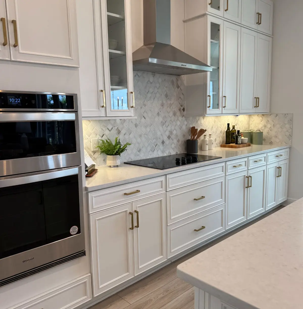White kitchen with glass front upper cabinets, brass hardware, marble diamond pattern backsplash, stainless range hood over a black cooktop, Whirlpool double wall ovens, quartz counters, and wood look tile floors.