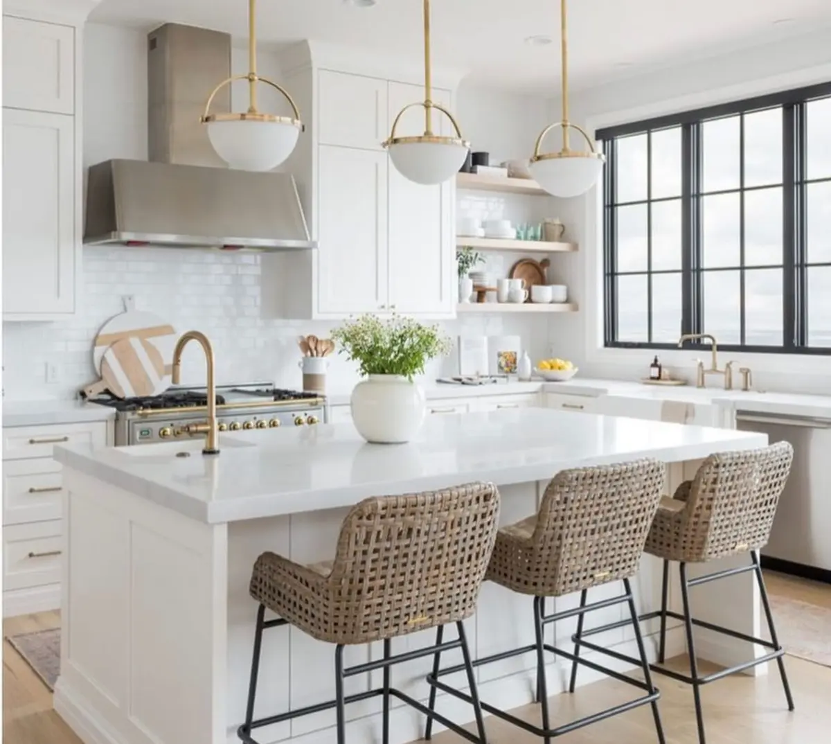 Bright white kitchen with a large quartz island, three woven barstools with black metal legs, brass pendant lights, brass faucets, stainless range hood, white subway tile backsplash, light wood open shelving, and black framed windows.