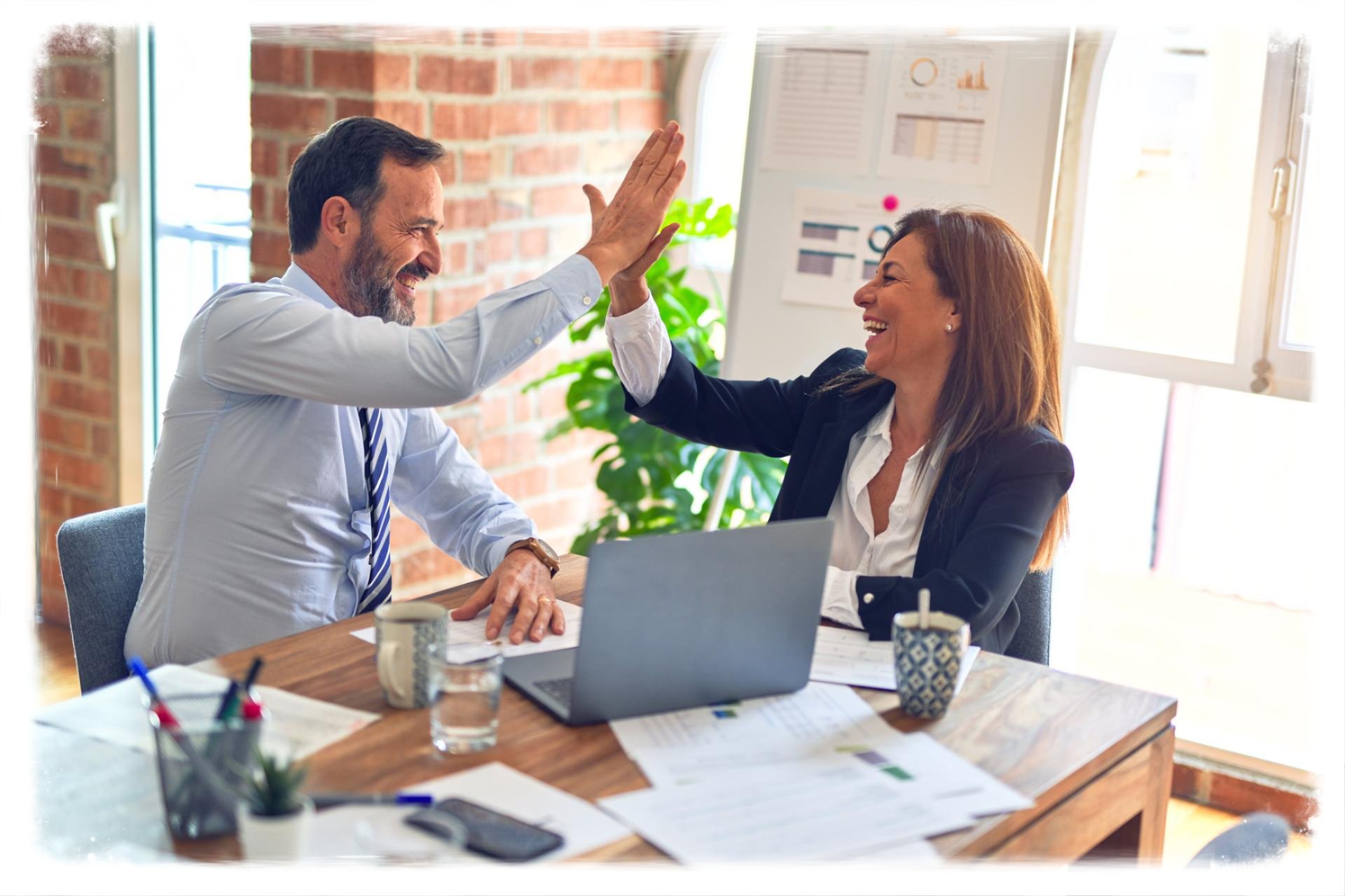 Two professionals smiling and high-fiving across a desk in a bright office, with a laptop, papers, and a plant visible, in a landscape composition with about a 1.62:1 aspect ratio.