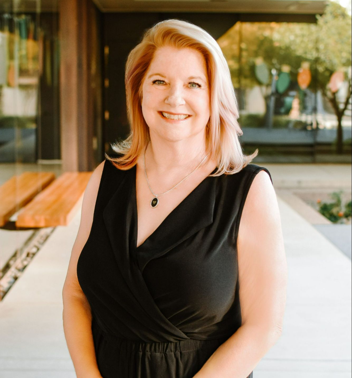 A smiling blonde woman in a black sleeveless outfit standing outdoors beside a modern building with warm reflections and greenery in the background, portrait photo in a roughly 357 by 373 aspect ratio.