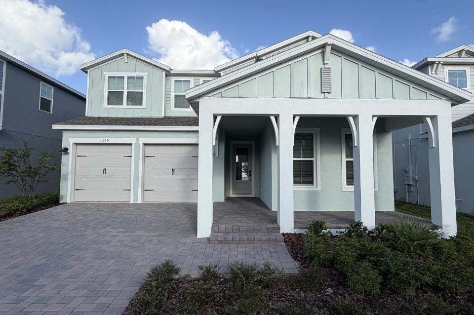 Pale green two-story home with a covered front porch and paver driveway under a blue sky